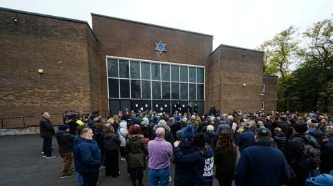 A few hundred people gather outside the synagogue a week after the attack. They are listening to speakers at an event in front of the brick building, which has a Star of David above central windows and glass doors.