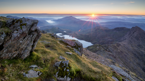 A vast, mountainous landscape is pictured from a summit looking down at lakes, clouds and grassland at sunrise.