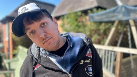 A young man in a baseball cap with black hooded jacket and blue shirt sat in a garden