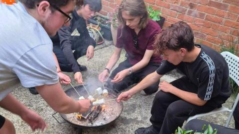 Four young teenage boys are toasting marshmallows on sticks round a fire bowl in a courtyard.