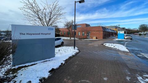 The outside of a hospital building with a view of the side of a car park. A sign that says The Haywood Hospital is at the forefront of the image. There is snow on the ground. 