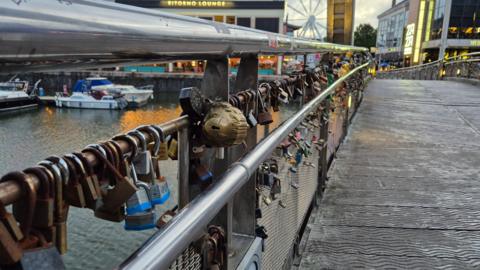 A metal footbridge over a harbour. The bridge is covered in padlocks. Boats are visible on the water and the lights of bars and restaurants can be seen in the background.