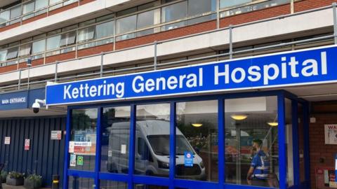 A blue and white sign that reads "Kettering General Hospital" above a glass-panelled door, with a security guard standing in a hallway. 