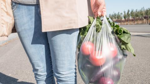 A woman carrying a clear plastic bag containing vegetables. Only their hands, legs and the bag can be seen in the frame. 