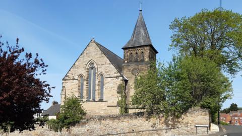 A honey-coloured Gothic Revival style stone church with tall pointed arched windows sits behind an old stone boundary wall with mature trees around it. Its square tower capped by a pyramidal roof is illuminated by the sunshine and stands out against the blue sky.