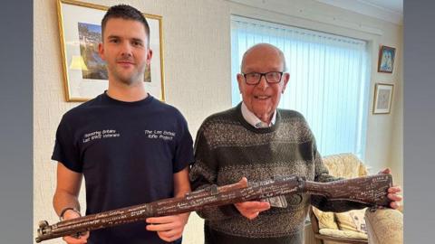 Jay Hawkins in a blue t-shirt saying the Lee Enfield Rifle Project standing beside Roy Hayward wearing a striped greeny-brown jumper over a white shirt both holding a Lee-Enfield Rifle between them