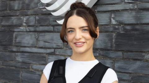 Olivia Browne yn sefyll o flaen wal y Senedd, yn gwisgo fest rhedeg