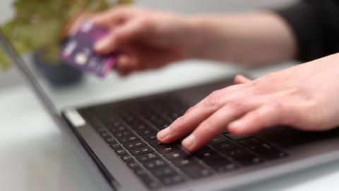 A woman's hands typing on a laptop placed on a white table with her left hand, and holding a purple bank card with her right hand. There is a plant in a pot next to the laptop.