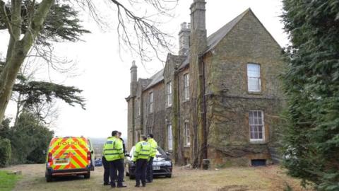 Exterior of two-storey stone-built house, with white window frames. Vegetation is growing up the walls. There are several chimneys above the front facade. A group of police officers, wearing yellow hi-vis police vests, are standing outside next to a car and a police van in  gravel drive. There is a tall tree to the left, and a shorter one to the right. 