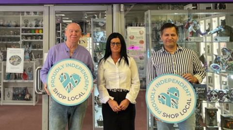 Two men and a woman stand outside a shop. The shop has shelves we can see showing metal figures, sculptures. The two men hold circular signs with the phrase "Proudly Independent - Shop Local" on them. The man on the left is bald with a purple shirt and jeans. The woman in the centre wears glasses, has dark hair past her shoulders and wears a white blouse and a black skirt. The man on the right has a black and white striped shirt, jeans and short dark hair