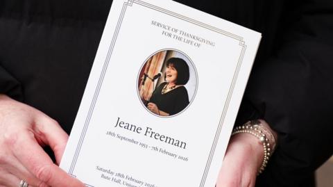 Hands holding an order of service which has a photo of Jeane Freeman on the front