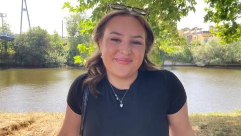 A woman with brown hair and sunglasses on her head. She is wearing a blue-black t-shirt and a silver heart necklace. She is standing in front of the bank, beyond which is the River Severn, with a footbridge visible on the left