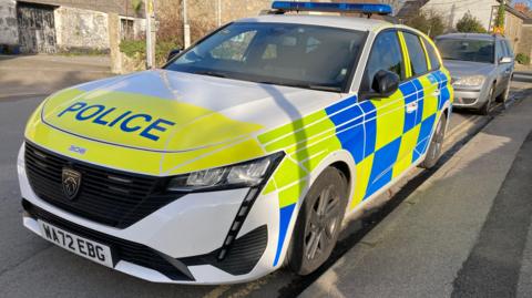 A white, blue and yellow Devon and Cornwall Police car is parked on a street. A grey Ford saloon car is parked behind it on double yellow lines. An old garage or barn building is in the background along with a row of houses. No-one is sitting in the police car.