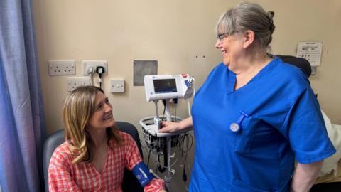 Patient, Hannah Robinson, wearing a red and white checked dress sitting in a hospital room smiling at a nurse standing in front of her, wearing a blue tunic and taking her blood pressure