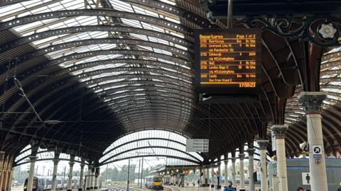 York train station from platform 3 with a timetable board listing the times of incoming trains. In the background is a train arriving into the station. 