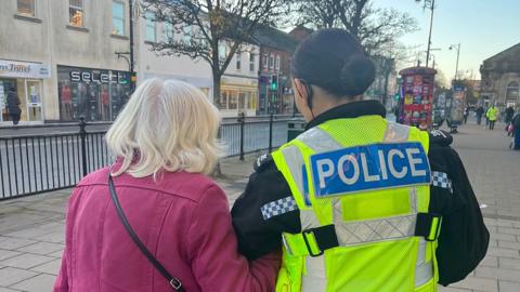 Rear view of Joan with white hair and wearing a pink jacket and PC Nicholson with dark hair in uniform walking to the bank to return the money