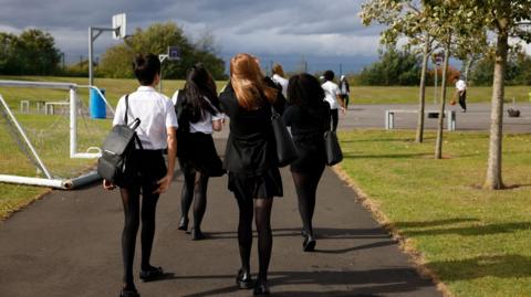 A stock image of girls and boys, taken behind them, wearing secondary school uniform and carrying backpacks while walking into a basketball court at a park.