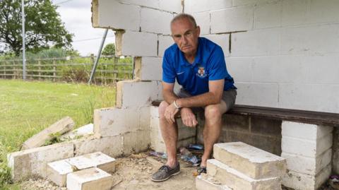 Len Keating sits on a bench inside a damaged concrete structure, surrounded by rubble and broken blocks. he is an older man with grey hair wearing sports kit. He wears a blue shirt and shorts and has a serious expression on his face. Greenery is visible through the gaps in the crumbling walls.