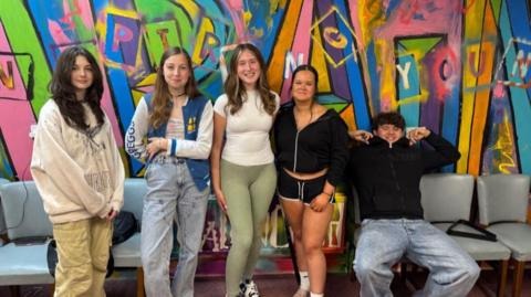 Yasmin Betteridge poses for a photo with two young people either side of her. Yasmin is wearing a white t shirt and light green leggings. Two teenage girls stand to her right and another stands to her left, a young man is sitting down at the end and smiling.  In the background is multicoloured graffiti art. 