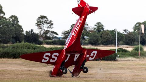 A red DH.88 Comet plane has its nose in the ground with brown soil being churned up by the propellers, which are bending in the ground