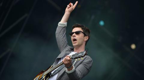 Kelly Jones of Stereophonics on stage. He has short dark hair, wears sunglasses, and is wearing a white t-shirt and grey blazer. He is holding a guitar and his hand is a aloft, pointing to the sky.