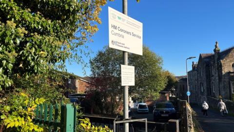 A general view of the entrance to the coroner's office in Cockermouth. There is a sign that says 'HM Coroners Service Cumbria' outside the entrance to a car park that leads to a low building.
