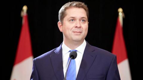 Andrew Scheer is photographed speaking at a podium. He is wearing a navy blue suit and a light blue tie. Behind him are two Canadian flags. He has short light brown hair and blue eyes.