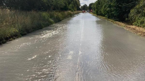 A road pictured from the middle of the carriageway, disappearing into the distance. It is completely underwater and you can barely make out the white lines. It is hedged by verges and trees on both sides and in the distance is a series of vehicles. 