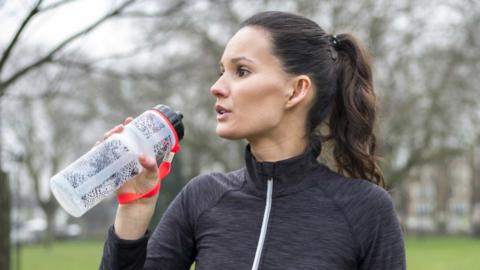 A woman wearing exercise clothes, standing in a park, and holding a sports bottle. 