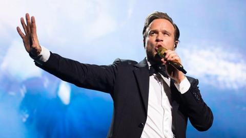 Olly Murs dressed in a black suit and white shirt sings into a mic during one of his concerts 