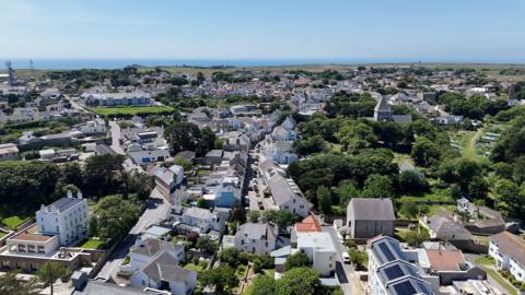 A picture of Alderney taken from above. There is a number of houses pictured with trees surrounding it.