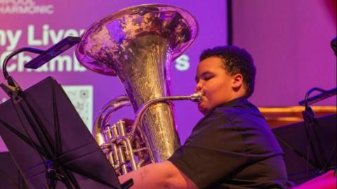 A boy is playing a tuba on stage at the Liverpool Philharmonic. He has short black hair and is wearing a black shirt.