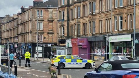 A street of tenement buildings - shops on the ground floor, flats above. A police car is parked in the middle of the road, and there is police tape across the full width of the street. A police officer in a hat and high-vis jacket is standing the road. 