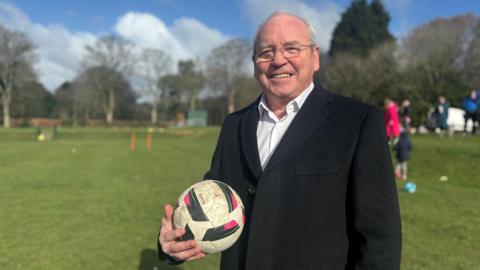 A man in his 60s, clean-shaven, wearing glasses and with thinning grey hair, stands on a grass field in a park. He is wearing a black blazer over a white shirt and holds a football in his right hand as he smiles for the camera.