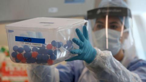 An NHS worker is holding a box of coronavirus tests. She is wearing a visor, gloves and an apron. You cannot see her face because she is also wearing a mask.