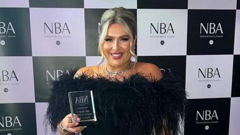 Charlotte Bates with blond hair worn up wearing an evening black velvet dress with black feathers on the collar. She is holding up a glass trophy she won at te National Beauty Awards.
