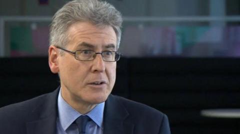 A man with grey hair and glasses in a dark suit and tie