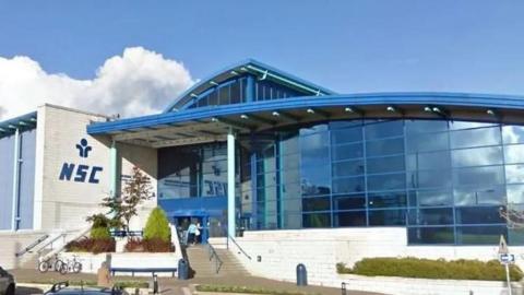 The outside of the National Sport Centre, which is a blue glass-fronted building with steps leading up to it. A white brick wall on the left hand side has the letters NSC on it in blue.