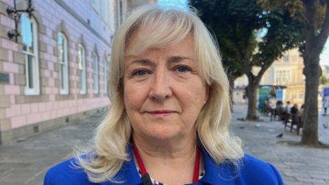Deputy Karen Wilson is wearing a blue blazer and has blonde hair. She is stood outside the States Assembly building. 