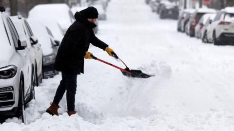 A man rugged up in heavy winter clothing uses a tool to clear snow packed into a shovel he is using to clear part of a road near a line of parked cars, all of which have snow over their roofs and windscreens.