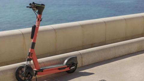 An orange e-scooter parked on a pavement and resting against a concrete sea wall. Beyond the wall is blue sea. 