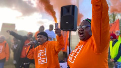 Protesters wearing orange jumpers light flares and raise their arms. Orange smoke from the flares is rising into the sky. One protester wearing a black wooly hat is speaking into a microphone and there is a speaker behind them. The other protester, on the right-hand side, has black glasses and a black wooly hat with coloured trim.