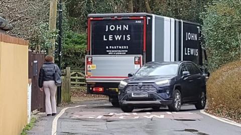 A picture of Walton Road in the village of Wavendon near Milton Keynes. You can see a woman trying to walk on a narrow path next to a road, where a John Lewis delivery van is trying to drive past a car.