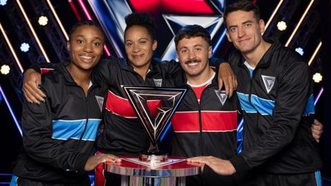 Four people in tracksuits around the Gladiators trophy
