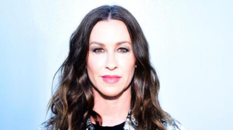Alanis Morissette stands against a light blue background. She has long wavy dark hair and is wearing a black and white collared shirt. 