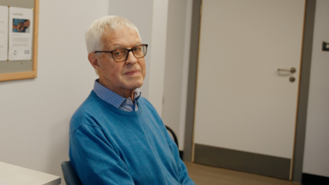 Howard Bartlett is sitting on a chair at a GP practice. He is wearing a blue sweater over a checked shirt. The room around him has white walls.