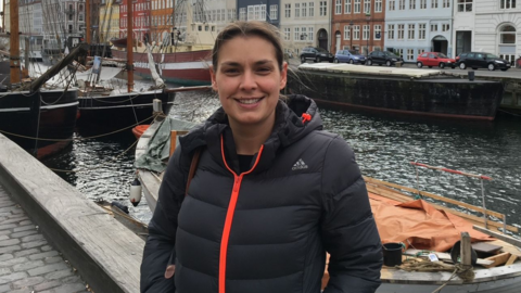 A woman with brown hair tied-up is wearing a black Adidas raincoat with an orange zip and smiling. She appears to be standing on a harbourside with boats behind her and a row of colourful houses.