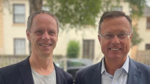 Sebastian Warrack and Robin Smith are both wearing blue jackets and smiling as they stand in a street outside Jersey Opera House with a pale cream building behind them.