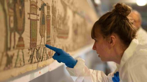 A woman in a sterile suit and gloves inspects the artwork with a pointed finger.