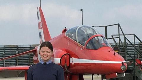 Holly stands in her navy blue cadet uniform smiling at the camera in front of a Red Arrow jet. Her dark hair has been tied back behind her head.